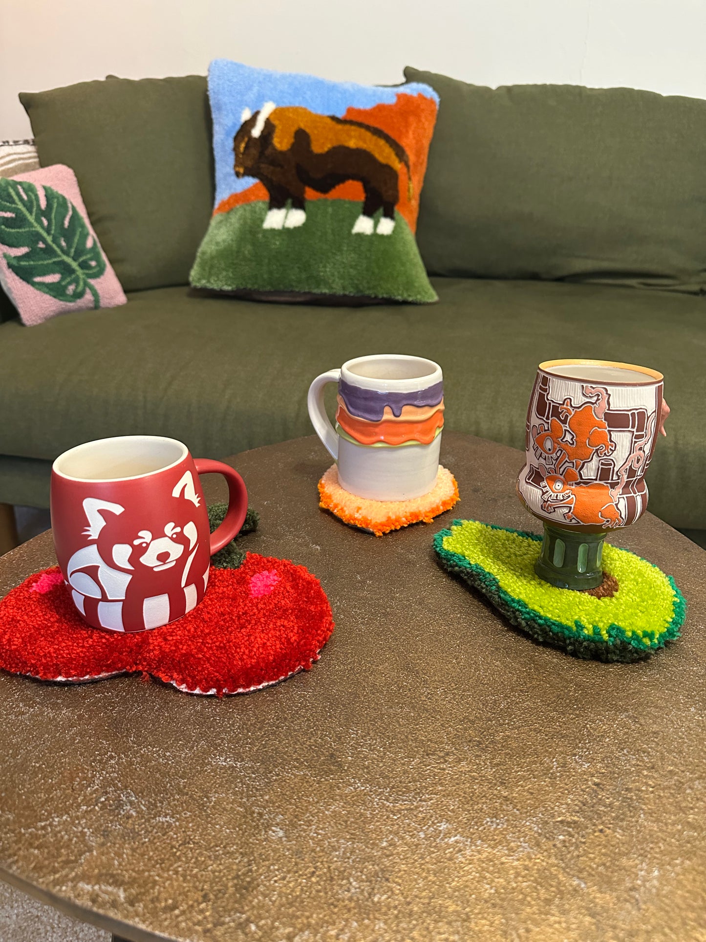 Three mugs on a table with decorative coasters and pillows on a couch in the background