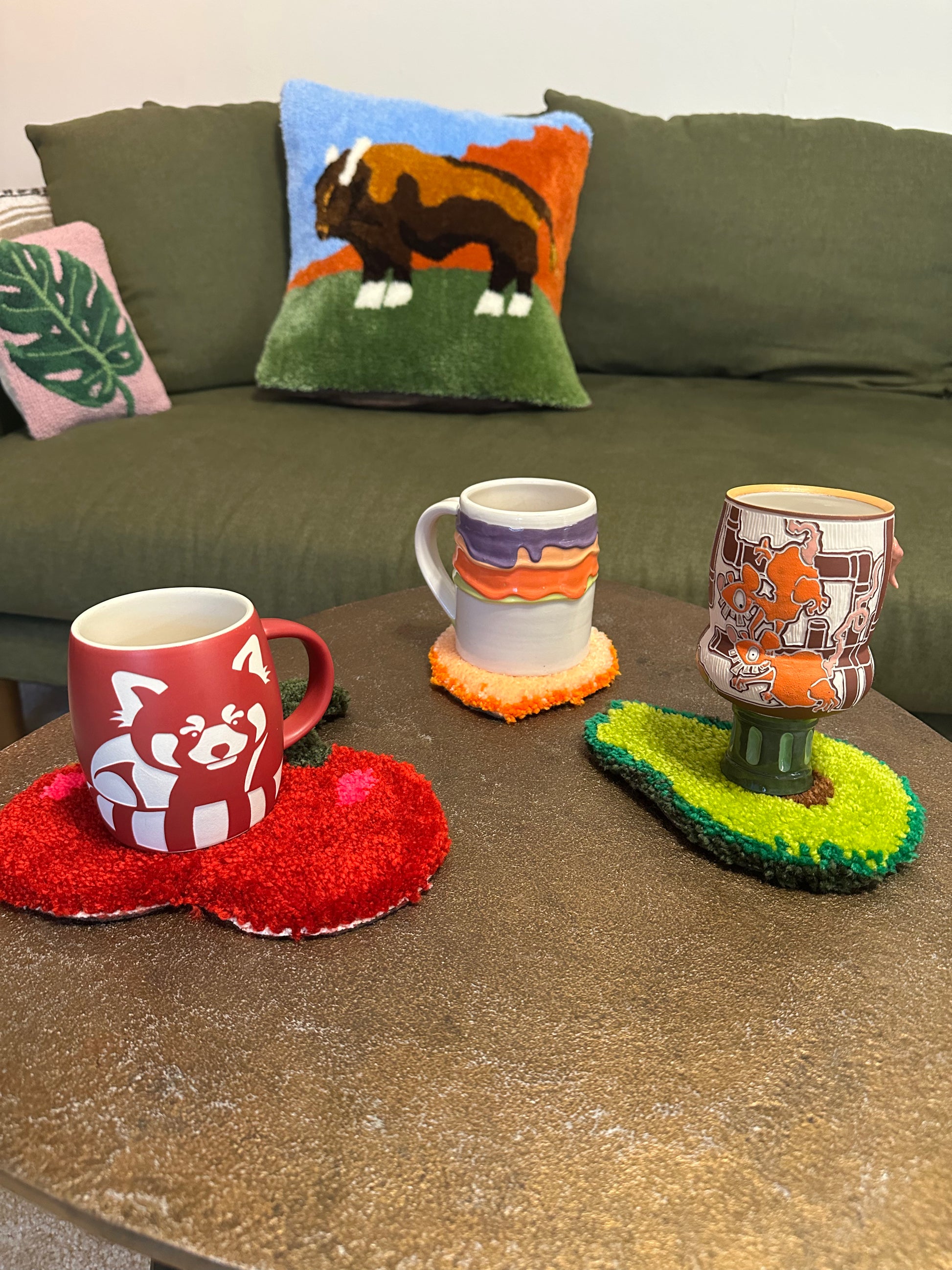 Three mugs on a table with decorative coasters and pillows on a couch in the background